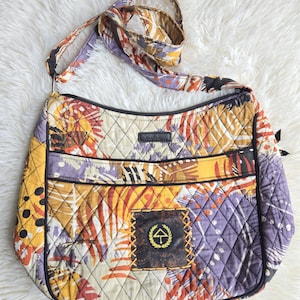 May include: A quilted crossbody bag featuring a vibrant abstract pattern in shades of yellow, orange, purple, and black. The bag includes an adjustable shoulder strap, a top zipper closure, a front exterior pocket, and a square patch with an umbrella logo.