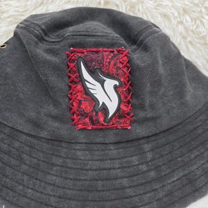 May include: A charcoal gray bucket hat featuring a rectangular patch stitched onto the front. The patch has a red paisley background with a white wing-shaped graphic in the center, secured by thick red cross-stitching around the edges.