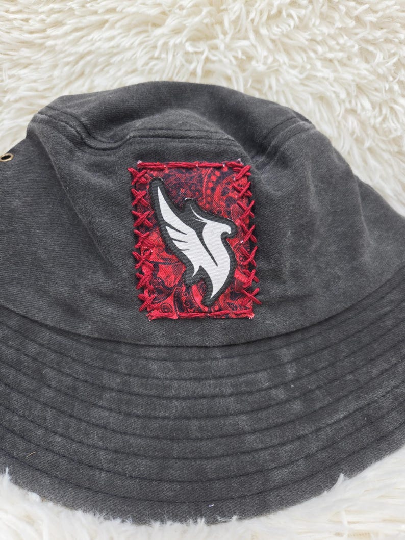 May include: A charcoal gray bucket hat featuring a rectangular patch stitched onto the front. The patch has a red paisley background with a white wing-shaped graphic in the center, secured by thick red cross-stitching around the edges.