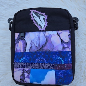 Puede incluir: Bolso bandolera negro con un diseño de patchwork en tonos morados y azules. El bolso tiene una cremallera en la parte superior y un emblema decorativo con una figura blanca con forma de pájaro. El bolso es rectangular y tiene correas negras ajustables.
