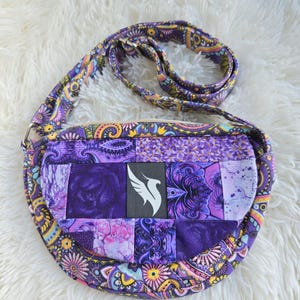 Puede incluir: Un bolso bandolera patchwork con una vibrante mezcla de estampados florales y paisley morados, rosas y amarillos. El bolso tiene una correa larga y un panel central con un logotipo de pájaro blanco sobre fondo negro. El bolso mide unos 20 cm de ancho.