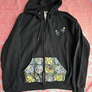 May include: A black zip-up hoodie featuring a hood with drawstrings and a front kangaroo pocket. The pocket displays a colourful stained-glass style pattern with various cartoon characters. Two small embroidered soot sprite figures appear on the chest.