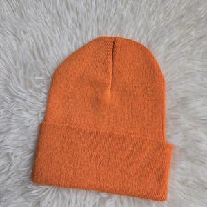 K&ouml;nnte beinhalten: Eine leuchtend orangefarbene Beanie-M&uuml;tze mit umgeschlagenem Rand. Die M&uuml;tze ist aus einem Strickmaterial gefertigt und hat eine einfarbige, leuchtend orange Farbe. Die M&uuml;tze liegt auf einer wei&szlig;en, flauschigen Oberfl&auml;che.