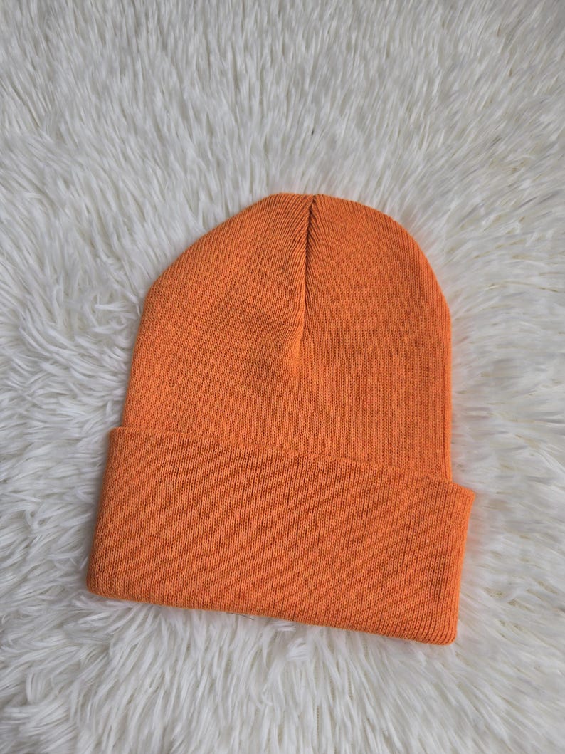 K&ouml;nnte beinhalten: Eine leuchtend orangefarbene Beanie-M&uuml;tze mit umgeschlagenem Rand. Die M&uuml;tze ist aus einem Strickmaterial gefertigt und hat eine einfarbige, leuchtend orange Farbe. Die M&uuml;tze liegt auf einer wei&szlig;en, flauschigen Oberfl&auml;che.