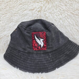 May include: A charcoal gray bucket hat featuring a rectangular patch on the front. The patch has a red and black paisley background with a white wing graphic stitched onto it. The hat includes a small metal eyelet on the side and stitched brim.