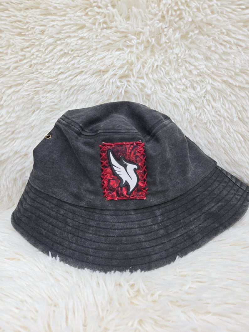 May include: A charcoal gray bucket hat featuring a rectangular patch on the front. The patch has a red and black paisley background with a white wing graphic stitched onto it. The hat includes a small metal eyelet on the side and stitched brim.