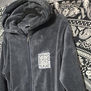 May include: A dark gray plush zip-up hoodie with a hood and long sleeves. The front features a rectangular patch with a white and black leopard print border surrounding a central graphic with the letters EG in a stylized font.