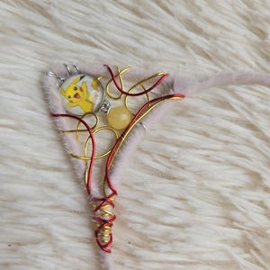 May include: A triangular cat ear accessory made of light pink faux fur, decorated with gold and red wire swirls, a small yellow bead, and a circular charm featuring a yellow Pikachu character illustration.