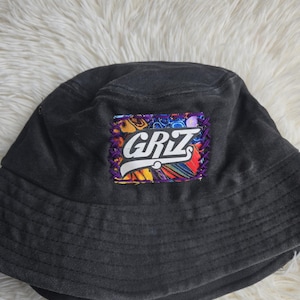 May include: A black bucket hat featuring a rectangular patch stitched onto the front. The patch displays the text GRIZ in white letters over a vibrant, multicoloured abstract background, secured with purple thread stitching around the edges.