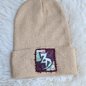 May include: A tan knit beanie featuring a rectangular fabric patch sewn onto the cuff. The patch consists of a patchwork background in shades of purple and floral patterns, with a black and white stylized graphic symbol stitched in the center.
