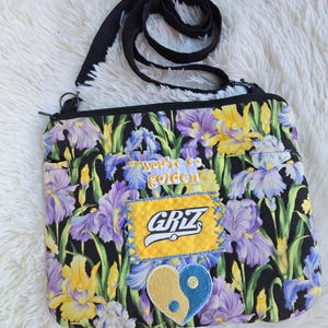 Könnte beinhalten: Eine rechteckige Umhängetasche mit schwarzem Stoffgrund und einem lebendigen Blumenmuster aus violetten und gelben Schwertlilien. Die Tasche verfügt über einen verstellbaren schwarzen Riemen, einen Reißverschluss oben und drei Aufnäher: den Schriftzug you're so golden, einen gelben GRIZ-Aufnäher und ein herzförmiges blau-gelbes Yin-Yang-Symbol.