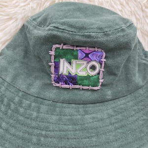 May include: A sage green bucket hat featuring a square patch on the front. The patch is divided into four quadrants with purple and green patterns, outlined in light purple stitching. The word INZO is printed in white letters on a black background.