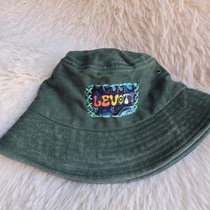 May include: A green bucket hat with a patch that reads "LEVO TY" in colorful lettering. The patch is stitched onto the hat with green thread. The hat is made of a soft material and has a wide brim.