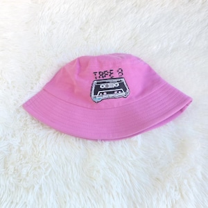 May include: A pink bucket hat featuring the text "TAPE 8" and a cassette tape design. The hat is made of a soft fabric and is shown on a white, fluffy surface.