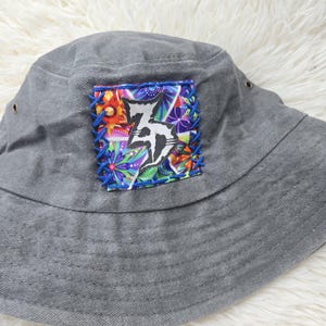 May include: A grey bucket hat featuring a square patch stitched onto the front. The patch displays a colorful, kaleidoscopic floral pattern with a black and white graphic symbol in the center, secured by thick blue thread lacing around the edges.