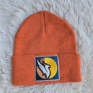 Può includere: Un berretto in maglia color arancione brillante con una toppa rettangolare sul risvolto. La toppa presenta uno sfondo quadrato blu scuro con un cerchio giallo e un grafico stilizzato di un'ala d'aquila bianca e nera al centro.