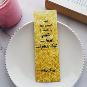 May include: A gold glitter bookmark with the quote "All the world is made of faith, and trust, and pixie dust" and the name "Peter Pan". The bookmark is on a white plate with a pink candle and a book in the background.