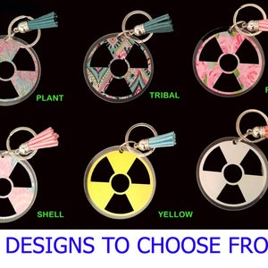 May include: Ten different designs of keychains with a black radiation symbol on a round, clear acrylic background. The keychains have different colored tassels and patterns on the back. The text "13 DESIGNS TO CHOOSE FROM!" is at the bottom of the image.