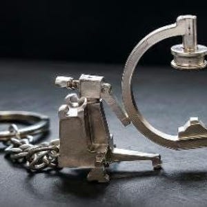 May include: A silver-toned metal keychain featuring a miniature C-arm medical imaging machine. The charm includes a curved arm, a central housing unit, and a circular ring attached by a short chain, all rendered in a polished metallic finish.