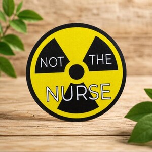 May include: A circular sticker featuring a yellow and black radiation hazard symbol. The text NOT THE NURSE is printed in white capital letters across the center of the design, with NOT THE positioned above the central circle and NURSE below.