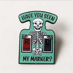 May include: A mint green enamel pin featuring a white skeleton holding two rectangular markers. The left marker is red with the letter R and the word RAD, while the right marker is black with the letter L and the word LEAD. Text reads HAVE YOU SEEN MY MARKER?