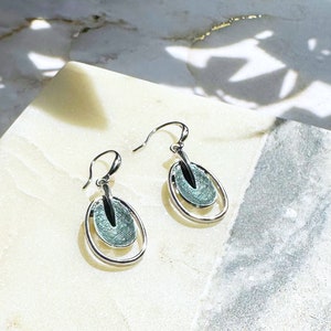A pair of rhodium plated open oval drop earrings with hook backings and a textured oval disc at the center painted in green enamel rest on a stone white and gray color block coaster underneath a plant casting shadows