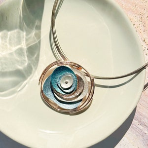 Adjustable Necklace with Brown Steel Wire chain and a round pendant made of 6 rose gold plated overlapping abstract textured discs painted in teal, blue, and gray enamel rests on a white plate on a purple and peach stone background
