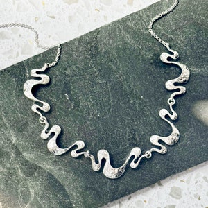 Adjustable Silver Necklace with a cable chain and five textured wave pendants connected by silver jump rings rests on a black stone tile on top of a white rocky background