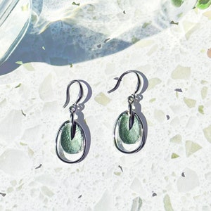 A pair of rhodium plated open oval drop earrings with hook backings and a textured oval disc at the center painted in green enamel rest on a textured white stone background next to a plant and a glass casting shadows