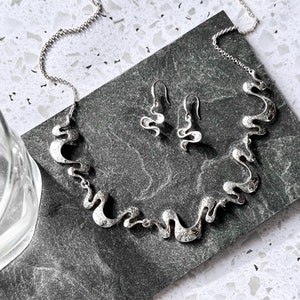 May include: A silver necklace and earrings set with a wavy, abstract design. The jewelry is on a dark gray background.