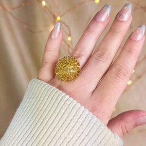 Puede incluir: Un anillo de oro con un diseño grande, redondo y puntiagudo. El anillo se lleva en el dedo índice de una mano con las uñas pintadas.