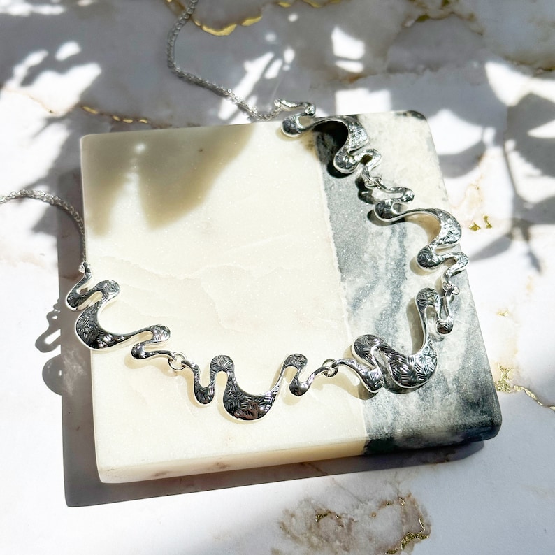 Adjustable Silver Necklace with a cable chain and five textured wave pendants connected by silver jump rings sits on a white and gray tile against a white marble background