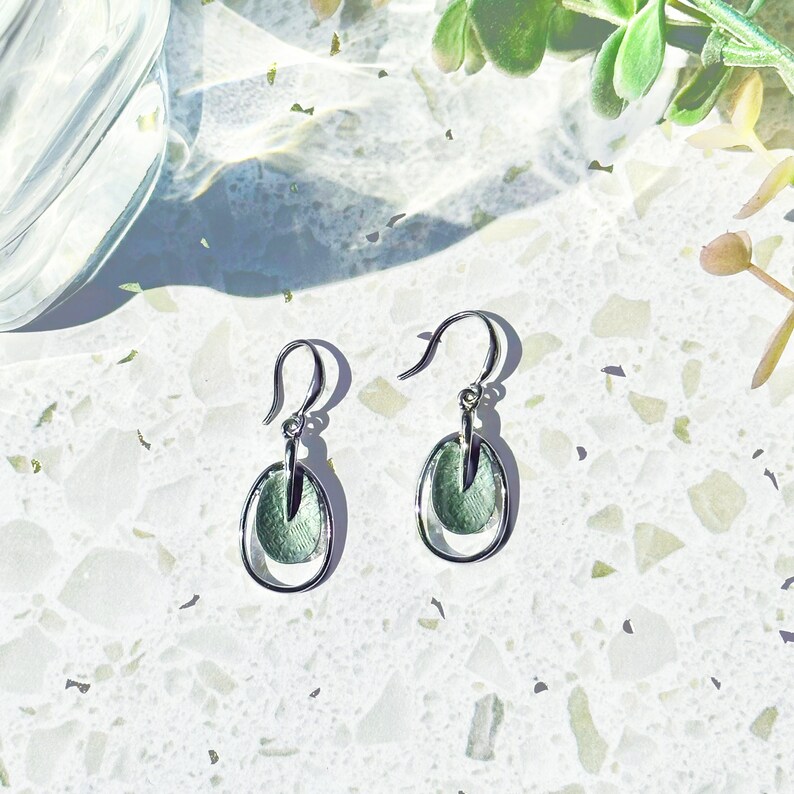 A pair of rhodium plated open oval drop earrings with hook backings and a textured oval disc at the center painted in green enamel rest on a textured white stone background next to a plant and a glass casting shadows