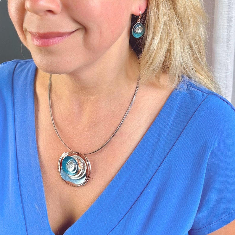 Necklace with Brown Steel Wire chain and a round pendant made of 6 rose gold plated overlapping abstract textured discs painted in teal, blue, and gray enamel with matching dangle earrings with hook backings worn by model wearing a blue blouse