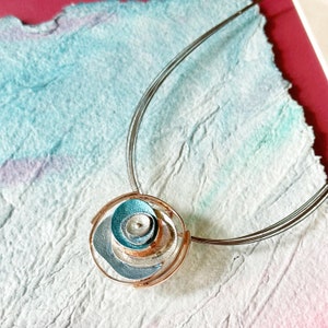Adjustable Necklace with Brown Steel Wire chain and a round pendant made of 6 rose gold plated overlapping abstract textured discs painted in teal, blue, and gray enamel rests on a pastel blue and purple painted cloth