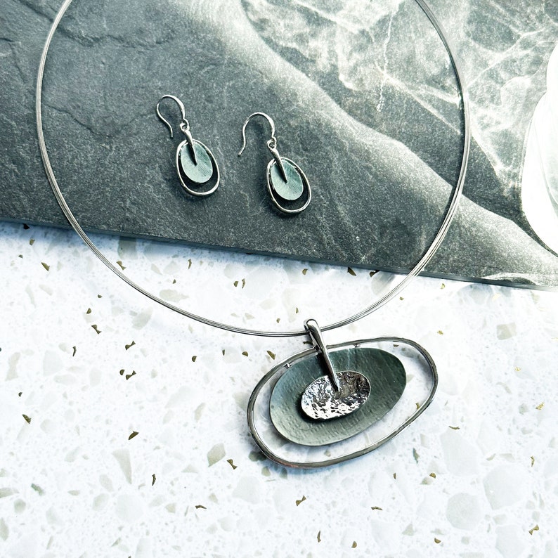 A pair of rhodium plated open oval drop earrings with hook backings and a textured oval disc at the center painted in green enamel rest on a textured gray stone tile on a white stone background with a matching necklace on a steel wire chain