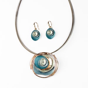 Adjustable Necklace with Brown Steel Wire chain and a round pendant made of 6 rose gold plated overlapping abstract textured discs painted in teal, blue, and gray enamel with matching dangle earrings with hook backings against a white background