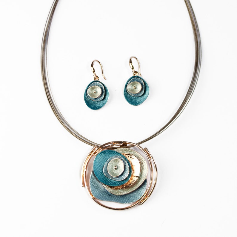 Adjustable Necklace with Brown Steel Wire chain and a round pendant made of 6 rose gold plated overlapping abstract textured discs painted in teal, blue, and gray enamel with matching dangle earrings with hook backings against a white background