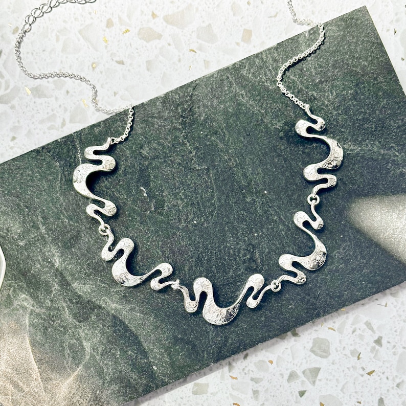 Adjustable Silver Necklace with a cable chain and five textured wave pendants connected by silver jump rings rests on a black stone tile on top of a white rocky background