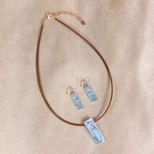 May include: A jewelry set featuring a necklace and matching earrings. The necklace has a brown cord and a rectangular pendant with a floral design. The earrings are also rectangular with a similar floral pattern. The metal accents are rose gold.