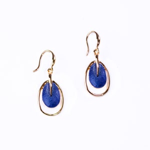 A pair of rose gold plated open oval drop earrings with hook backings and a textured oval disc at the center painted in blue enamel rest against a white background.