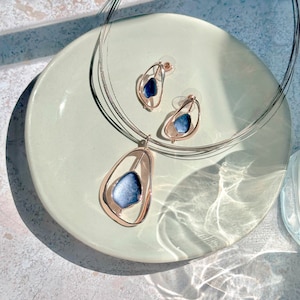 Adjustable Necklace with a brown steel wire chain and an open Pear shape rose gold pendant with a floating textured disc center painted in blue enamel with matching earrings rest on a white plate on a textured stone background with a glass