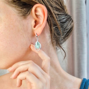A pair of rhodium plated open oval drop earrings with hook backings and a textured oval disc at the center painted in green enamel are worn by a model with her head turned to the side and her hand by her jawline