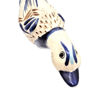Könnte beinhalten: Eine Keramik-Entenfigur mit cremefarbenem Körper, verziert mit braunen und blauen Malereien. Der Entenkopf ist nach unten geneigt und zeigt dunkle Augen und einen blauen Schnabel. Die Figur hat eine strukturierte Oberfläche.