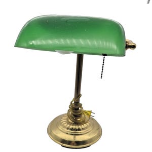 May include: A banker's lamp featuring a translucent emerald green glass shade, a brass-toned metal base, and a fluted central column. A black pull chain hangs from the shade, and a coiled power cord with a yellow plug rests on the base.