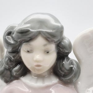 May include: A porcelain angel figurine with grey hair and a pink dress. The angel has a pale face with delicate features and downcast eyes. White wings are partially visible. The figurine is a decorative item.