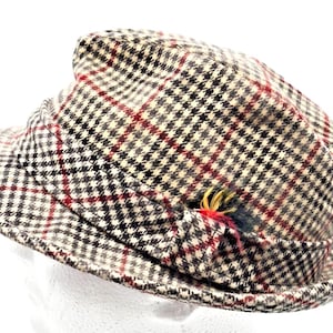 May include: A fedora hat featuring a beige, black, and brown houndstooth pattern with thin red overcheck lines. The hat includes a matching fabric band around the crown and a small decorative feather accent tucked into the side of the band.