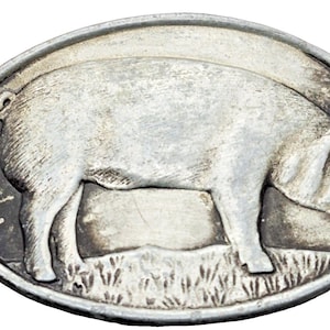 May include: Oval-shaped silver-toned metal belt buckle featuring a detailed relief of a pig standing on grass. The pig is facing right, with its curly tail visible. The buckle has a slightly aged appearance.