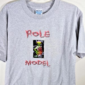 May include: A light gray Gildan t-shirt with the words "Role Model" in red, stylized lettering. The shirt features a graphic of a green alien holding a basketball. The alien is inside a black rectangle.