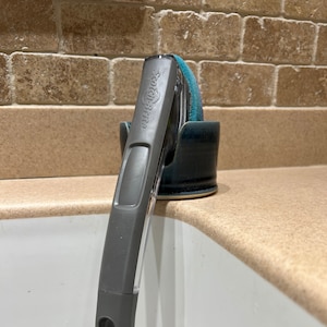 May include: A gray Scotch-Brite dish wand with a clear handle section and a teal scrubbing sponge head, resting upright in a dark blue ceramic holder. The handle features a rectangular cutout at the base and the brand name embossed on the side.
